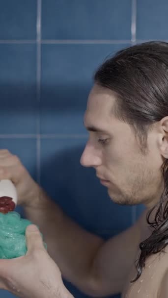 Joven hombre de pelo largo vertiendo un poco de jabón de baño en una esponja de baño en la ducha - Primer plano - Vídeo vertical - Metraje, vídeo