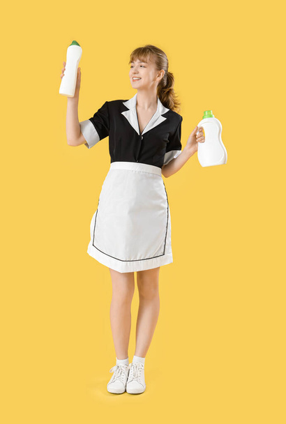 Junges Zimmermädchen mit Waschmittelflaschen auf gelbem Hintergrund - Foto, Bild