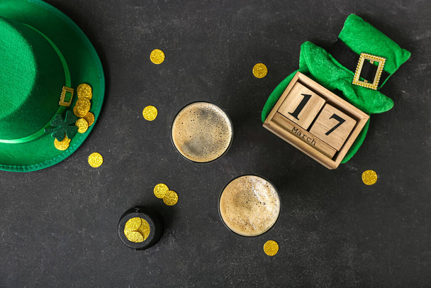 Glasses of dark beer with leprechaun hats, pot of golden coins and calendar for St. Patrick's Day celebration on black grunge background - Photo, Image