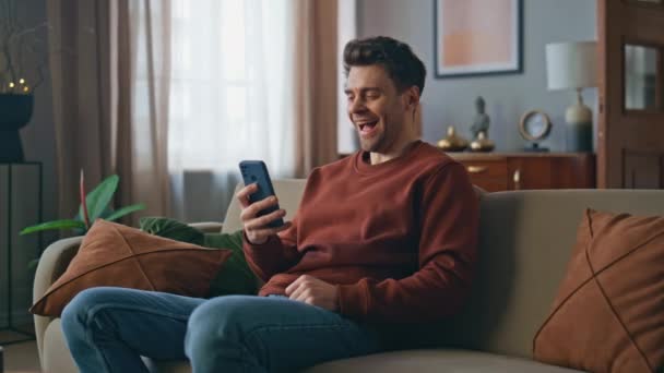 Смеющийся парень смотрит смешные видео со смартфона на диване. Счастливый улыбчивый мужчина просматривает ленту новостей, наслаждаясь развлекательным контентом на мобильном телефоне. Веселый мужчина смотрит социальные сети на мобильном телефоне - Кадры, видео