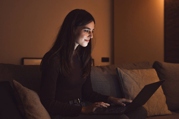 Zbliżenie do jednej młodej szczęśliwej kobiety uśmiechającej się i korzystającej z laptopa w domu siedząc na kanapie pracując i ucząc się samotnie w nocy. Millennial dziewczyna surfing w sieci kryty z ekranem świetlnym na twarzy relaks na kanapie. - Zdjęcie, obraz