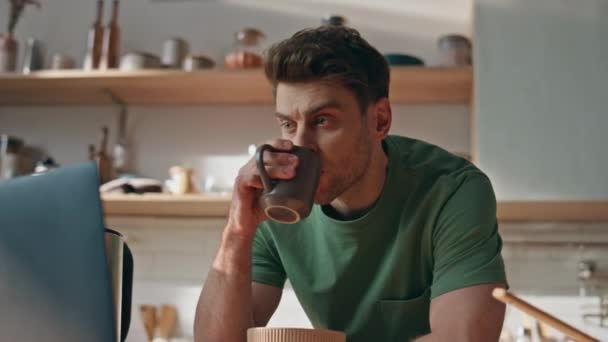 Nyugodt hipszter kávézik filmet néz a konyhában közelről. Fiatal barna férfi néz laptop képernyő kezében forró tea csésze a modern belső térben. Nyugodt fickó, aki otthon reggelizik. - Felvétel, videó