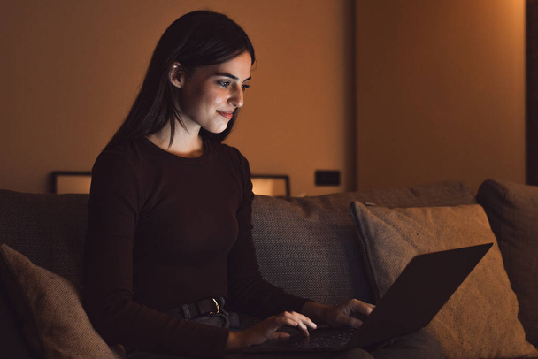 Großaufnahme einer jungen, fröhlichen Frau, die lächelt und zu Hause einen Laptop benutzt, auf dem Sofa sitzt, arbeitet und spät in der Nacht allein lernt. Millennial-Mädchen surft drinnen im Netz mit Lichtbildschirm im Gesicht entspannt sich auf der Couch. - Foto, Bild