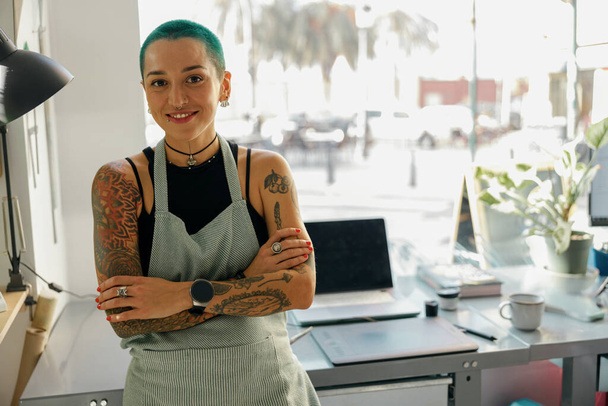 Glimlachende vrouwelijke ontwerper in schort staat in de buurt van bureau met laptop in eigen atelier en kijkt camera - Foto, afbeelding