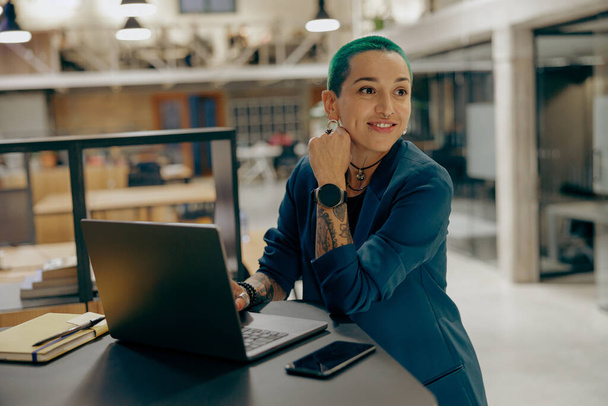 Усміхнена татуйована фрілансерка з зеленим коротким волоссям, що працює на ноутбуці, сидячи в офісі - Фото, зображення