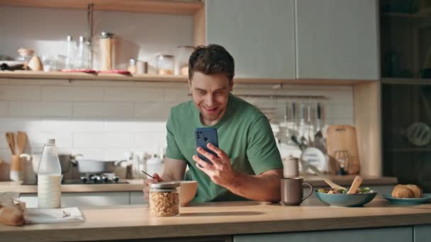 Feliz hombre leyendo el mensaje del teléfono celular en el primer plano del mostrador de la cocina. Guapo millennial chico disfrutando de almuerzo recibir una buena notificación en el teléfono inteligente. Hombre de negocios relajado desayunando antes de trabajar solo - Metraje, vídeo