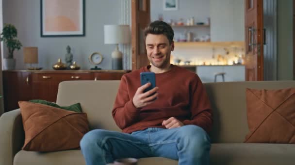 Fröhlicher Mann, der auf der heimischen Couch die sozialen Medien verfolgt. Glücklich entspannter Typ, der lachend auf lustige Online-Videos am Smartphone-Bildschirm schaut. Lächelnder Mann sitzt gemütlich auf Sofa mit Handy. - Filmmaterial, Video