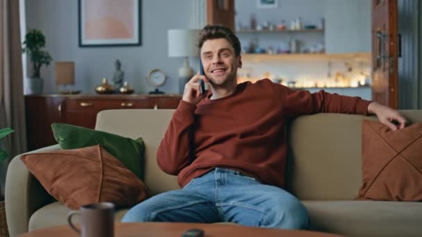 Rustende man communiceert smartphone in gezellige woonkamer bank. Een glimlachende knappe man die in het weekend nieuws deelt. Happy hipster ontspannen op de bank genieten van een aangenaam gesprek. - Video