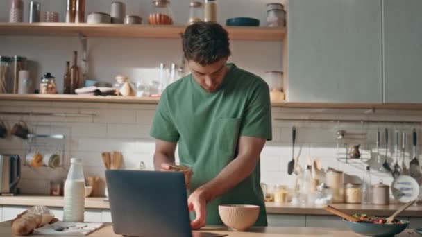 Vaření chlap vypadá notebook připravuje snídani v moderní kuchyni detailní up. Zapojený bruneta muž čtení zprávy v soukromém domě. Soustředěný pohledný muž kontroluje e-mail na počítači doma - Záběry, video