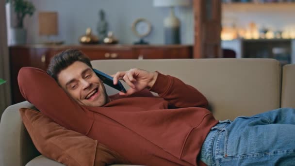 Boldog diák beszél mobiltelefon fekvő kényelmes kanapé közelről. Pozitív jóképű férfi élvezi a barátságos kommunikációt okostelefonnal otthon. Mosolygó fiatal szabadúszó beszél barátaival a hazai hétvégén. - Felvétel, videó