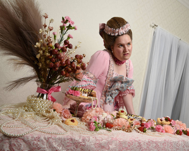 Primer plano retrato de modelo femenina linda con un vestido rosa opulento, el traje de una nobleza barroca francesa histórica. Comer comida deliciosa con pasteles, flores y joyas ricas - Foto, Imagen