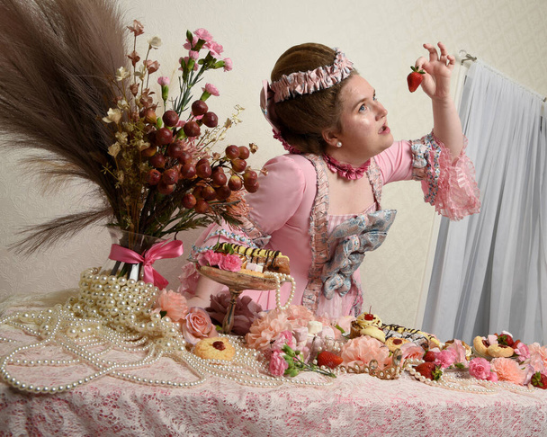 Close up portret uroczej modelki w przestronnej różowej sukni, kostium historycznej francuskiej szlachty barokowej. Jedzenie pobłażliwej uczty z ciasta, kwiaty i bogate klejnoty - Zdjęcie, obraz
