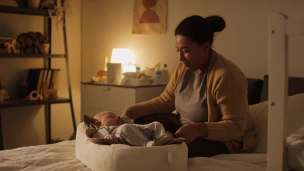 Pełne ujęcie młodej Afroamerykanki lub latynoskiej matki siedzącej w nocy w łóżku, tulącej swoje śpiące dziecko w kokonie, pochylającej się, podziwiającej i pieszczącej go z kochającym spojrzeniem - Materiał filmowy, wideo
