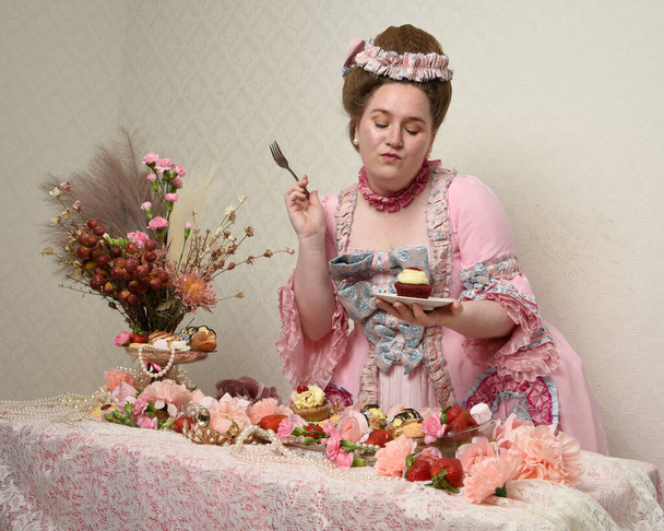 Close-up portret van schattig vrouwelijk model met een weelderige roze jurk, het kostuum van een historische Franse barokke adel. Het eten van taarten op een verwennend feest met zoete lekkernijen en rijk voedsel. - Foto, afbeelding