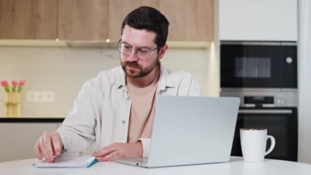 Modern bilgisayardaki web sitelerine göz atarken notlar alan yakışıklı bir adamın ön görüntüsü. Çevrimiçi üretken işlerde yetenekli genç serbest çalışan. Tasarımcı mutfağının arka planı. - Video, Çekim
