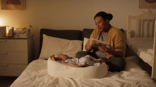 Volledig shot van Spaanse of Afro-Amerikaanse vrouw zitten naast pasgeboren zoon slapen in baby cocon, het nemen van kinderen verhaal boek en voorlezen van hardop sprookje voor zoete dromen - Video