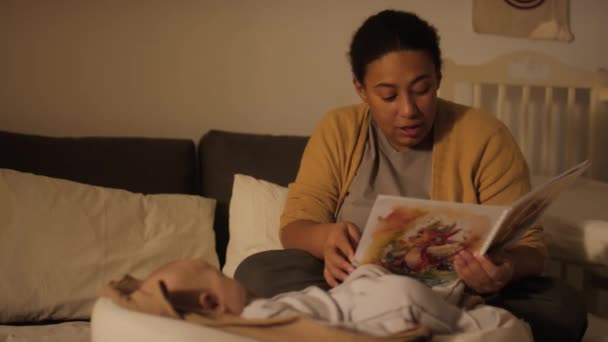 Medium full shot of young biracial woman sitting beside newborn son or daughter sleeping in baby cocoon and reading aloud fairy tale from colourful comic book about little dinosaurs - Footage, Video