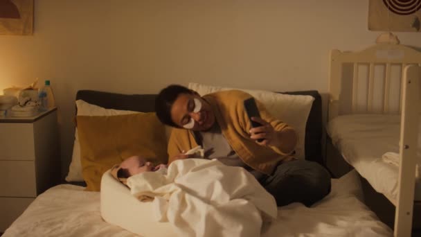 Plan complet de jeune femme afro-américaine ou hispanique vêtue de façon décontractée avec des patchs sous les yeux assis sur le lit à la maison, ayant un appel vidéo sur smartphone, souriant et posant avec bébé - Séquence, vidéo