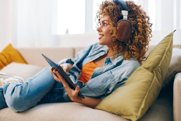 Boldog fiatal nő kezében egy tabletta PC és fejhallgató, olvasás és zenét hallgatni a kanapén otthon. - Fotó, kép