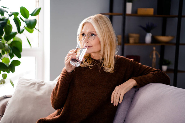 Photo of mature charming woman sitting sofa drinking fresh water vitamins on sick leave indoors. - Фото, зображення