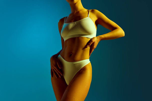Ausgeschnittenes Porträt einer durchtrainierten Frau in cremefarbener Unterwäsche in gelbem Neonlicht vor blauem Hintergrund. Konzept der Schönheitsbehandlungen, Diäten, weibliche Gesundheit, Weiblichkeit, Wellness-Verfahren. - Foto, Bild