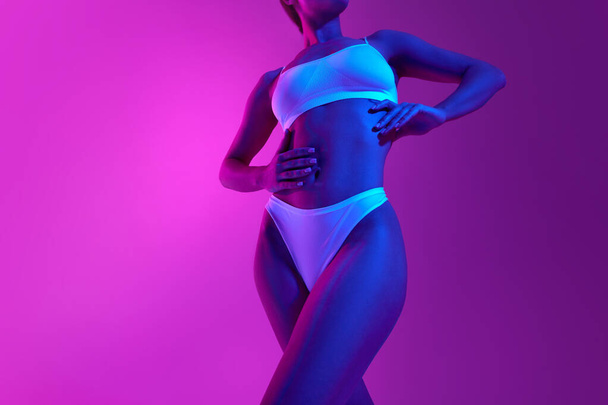 Manter o corpo tonificado. Imagem de close-up do corpo feminino magro, barriga em luz de néon azul contra fundo roxo gradiente. Conceito de tratamentos de beleza, dieta, saúde feminina, feminilidade, procedimentos de spa. - Foto, Imagem