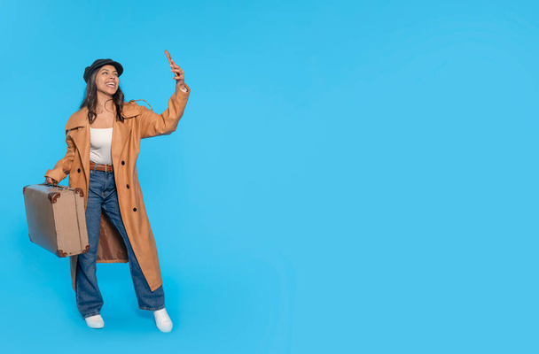 uma mulher de jeans, uma camiseta branca e um casaco castanho carregando uma mala e tirando uma selfie por telefone em um fundo azul. Pessoas felizes indo de férias, férias - Foto, Imagem