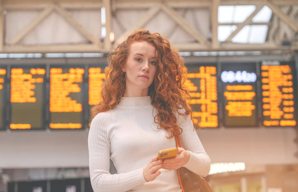an excited redhead female holding a smartphone while at the train station and the train is arriving. Safety travel concept - Photo, Image