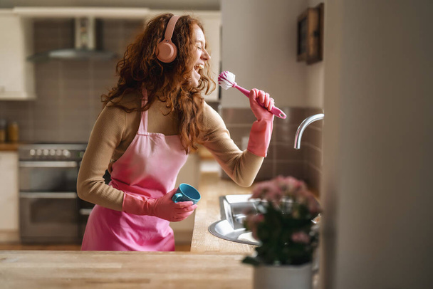 Руда жінка миє посуд на кухні і слухає музику за допомогою бездротових навушників, щаслива жіноча чистка пластини з миючим засобом, насолоджуючись виготовленням домашніх справ - Фото, зображення