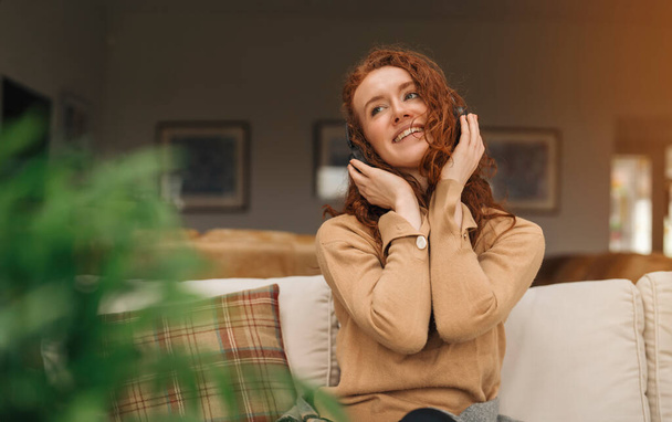 Ευτυχισμένος νεαρή κοκκινομάλλα γυναίκα σε καφέ πουκάμισο και χρησιμοποιώντας ένα κινητό τηλέφωνο, ακούγοντας αγαπημένα τραγούδια, audiobooks, podcast σε ακουστικά, ενώ κάθεται κοντά σε έναν καναπέ στο σπίτι - Φωτογραφία, εικόνα
