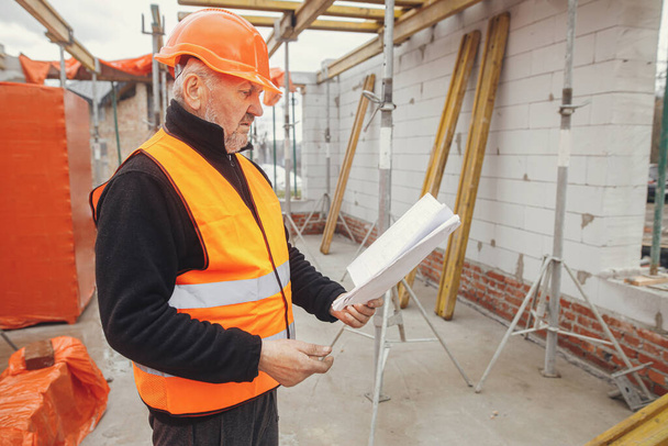 Senior man engineer or construction worker in hardhat looking at blueprints at building new modern house. Male architect or contractor checking plans at construction site. Copy space - Zdjęcie, obraz
