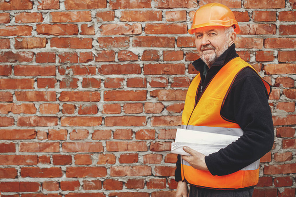 Senior male foreman in hardhat standing with blueprints at masonry brick wall at building new house. Portrait of happy man engineer or construction worker with plans at construction site - Photo, Image