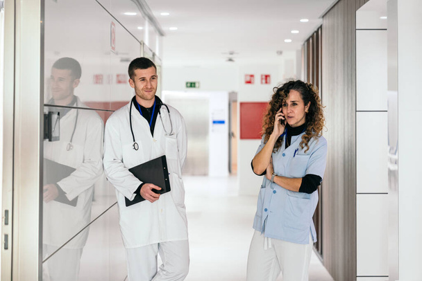 Verpleegster aan de telefoon terwijl de dokter aantekeningen bekijkt in een ziekenhuisgang. - Foto, afbeelding