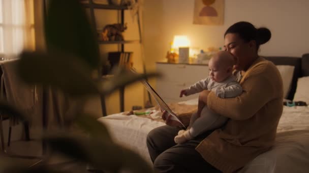 Mediana toma completa de la joven afroamericana sentada en la cama en casa, abrazando al bebé hijo en el regazo, sosteniendo un libro de cuentos de hadas, leyendo en voz alta y mirando fotos juntos - Metraje, vídeo