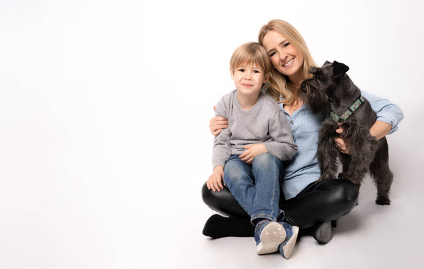 Mooie jonge moeder met kind jongen en puppy schnauzer hond zitten op de vloer op witte achtergrond - Foto, afbeelding