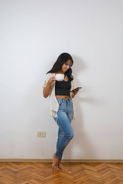 verticaal beeld, jong latijn brunette Argentijnse vrouw thuis staan drinken koffie leunend tegen de muur en met behulp van telefoon kijken sociale media, surfen op internet tijdens het nemen van pauze. - Foto, afbeelding