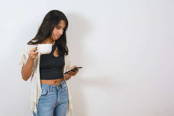 junge lateinische brünette Argentinierin steht zu Hause und trinkt Kaffee, lehnt sich an die Wand und benutzt das Handy, um soziale Medien zu sehen, surft während der Pause im Internet. - Foto, Bild