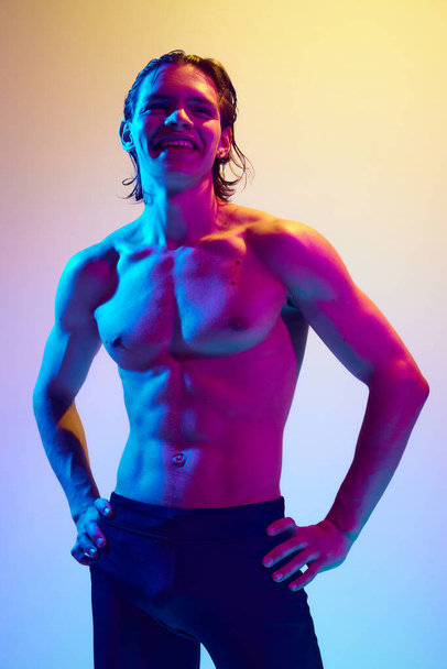 Un giovane uomo sorridente si tiene per mano sui fianchi dimostrando le sue curve del corpo perfette in una vibrante luce al neon su uno sfondo sfumato blu-rosa. Concetto di bellezza naturale persone, salute maschile, mascolinità. - Foto, immagini
