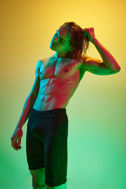 Retrato de jovem atleta atraente homem tocando seu cabelo molhado em luz de néon multicolorido contra fundo estúdio gradiente. Conceito de pessoas de beleza natural, fitness, saúde masculina, masculinidade. - Foto, Imagem