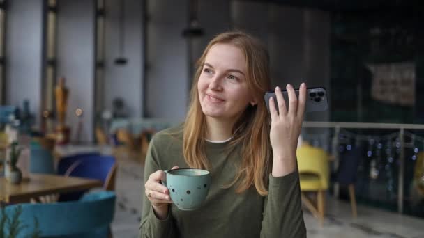 クラクフのトレンディな居心地の良いコーヒーショップカフェに座っている電話の声認識を使用してブロンドの女性の肖像画  - 映像、動画