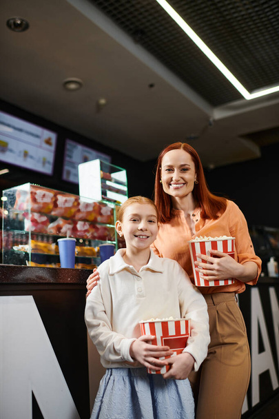 Kobieta stoi obok córki, trzymając dwa pudełka popcornu, ciesząc się rodzinną wycieczką do kina.. - Zdjęcie, obraz