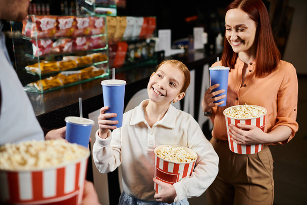 Onnellinen perhe seisoo yhdessä ympyrässä, pitäen ämpäreitä popcornia elokuvateatterissa. - Valokuva, kuva