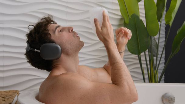 Calm tranquil Caucasian naked relaxed guy sitting in bathroom resting man relax in foamy bathtub blow foam enjoy audio in earphones male relaxing at home bath hotel listening music song in headphones - Foto, immagini