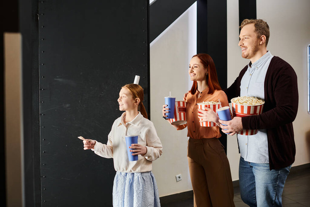 Μια ευτυχισμένη οικογένεια στέκεται κοντά, κρατώντας ποπ κορν σε ένα κινηματογραφικό σκηνικό, απολαμβάνοντας μια χαρούμενη στιγμή μαζί. - Φωτογραφία, εικόνα