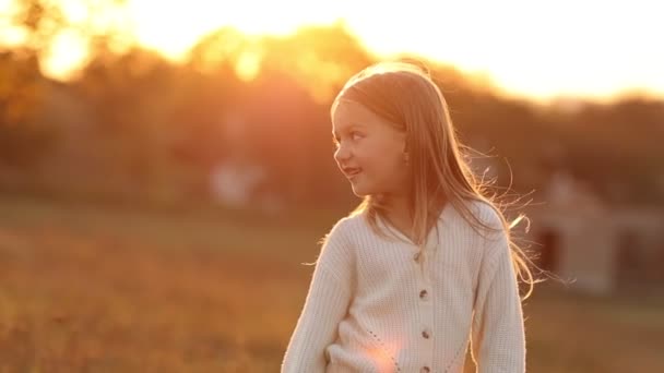 Journée de l'enfance. Joyeux petit enfant fille caucasienne 5-6 ans profite des vacances d'automne sur les lumières du coucher du soleil à la nature, regarde sur le côté avec des cheveux longs volant sur le vent. Activité de loisirs positive. - Séquence, vidéo