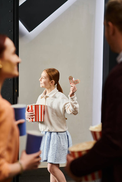 Dziewczyna z radością trzyma pudełko po popcornie i bilety, ciesząc się rodzinną nocą filmową w kinie. - Zdjęcie, obraz