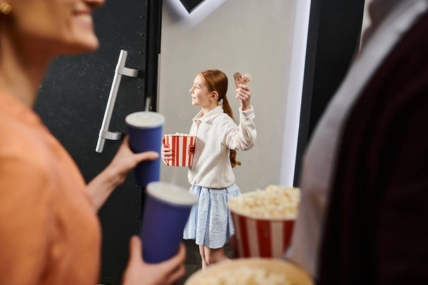 子供は喜んで映画館の家族の隣に立っている間ポップコーンのカップを握ります. - 写真・画像