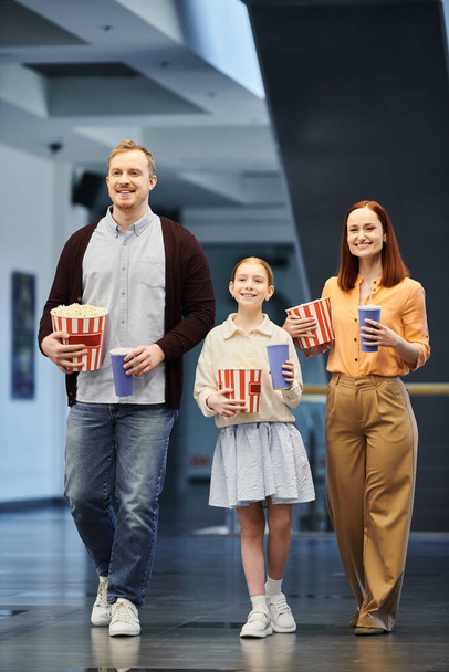 Mężczyzna, kobieta i dziecko chodzący szczęśliwie korytarzem w kinie, cieszący się dobrym spędzonym razem z rodziną. - Zdjęcie, obraz