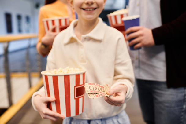 Mädchen hält freudig Eimer mit Popcorn und Eintrittskarten in einem Kino, umgeben von seiner glücklichen Familie. - Foto, Bild