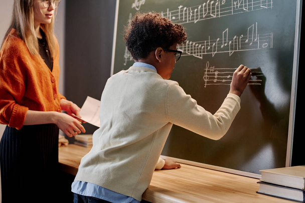 Menino afro-americano escrevendo notas quadro negro durante a aula de música, sua professora branca do sexo feminino que está ao lado dele - Foto, Imagem